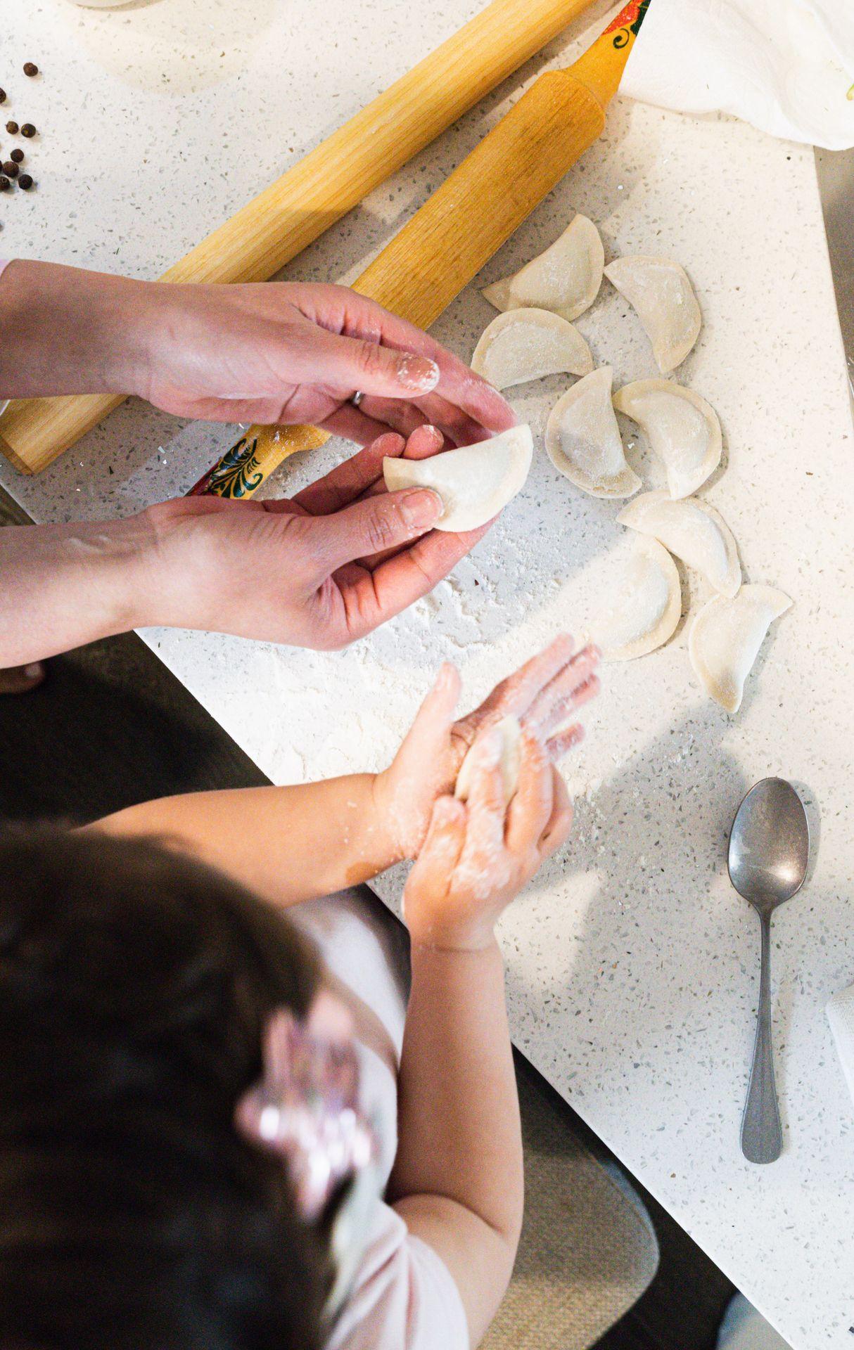 Child and adult hands preparing dumplings on a countertop with flour, rolling pin, and spoon.