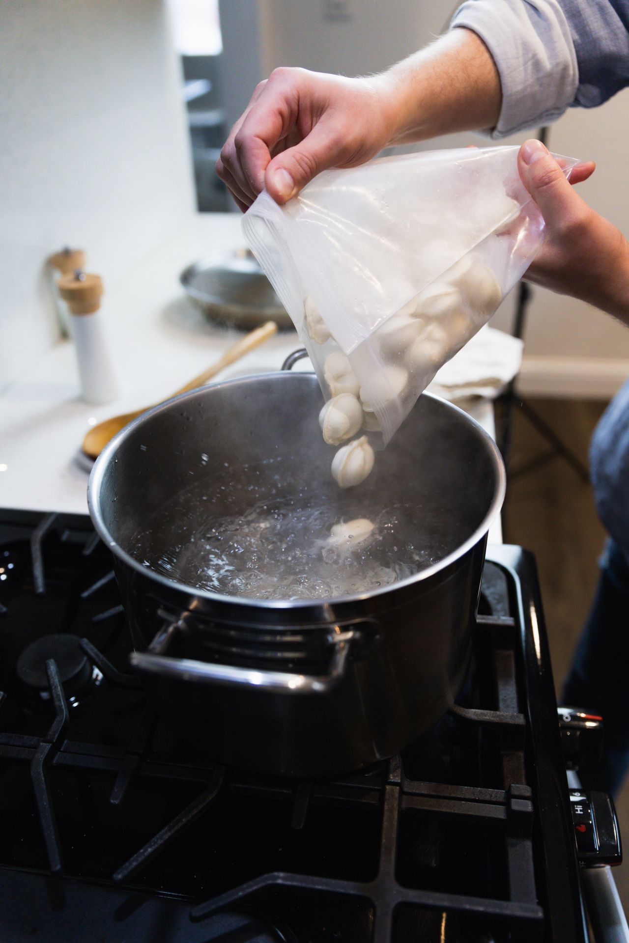 A person adding dumplings from a plastic bag into a pot of boiling water on a stove.